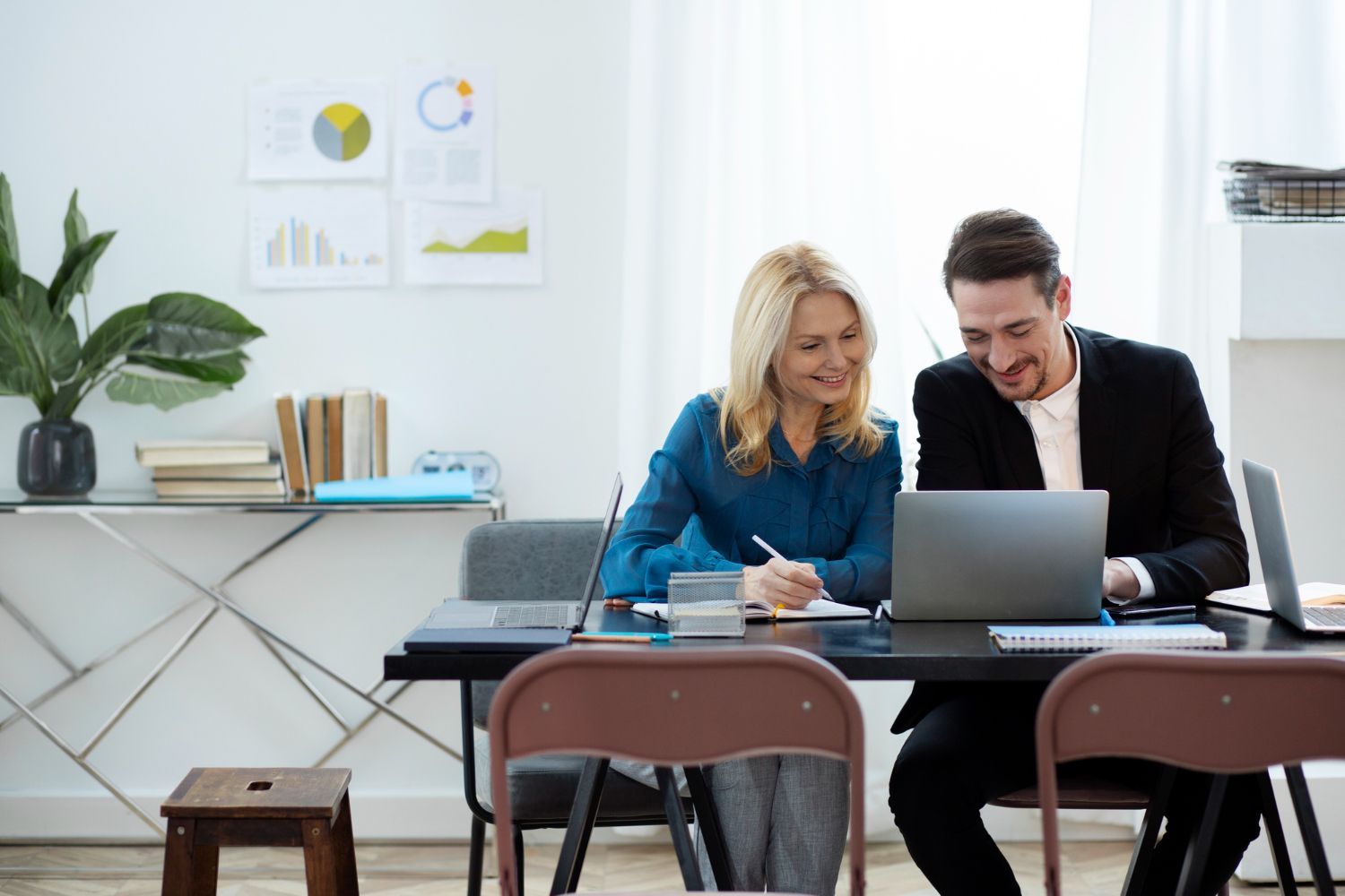 Two coworkers collaborating at a laptop in a bright office, with charts on the wall