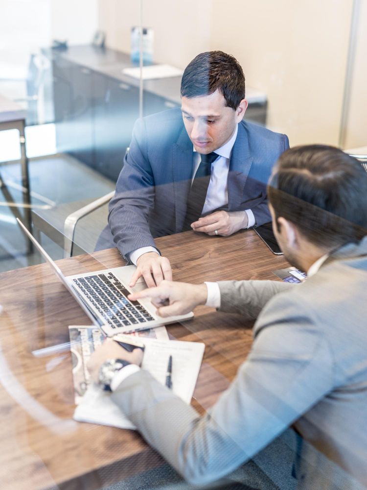 Two coworkers reviewing a laptop and documents at a conference table in a glass-walled office.