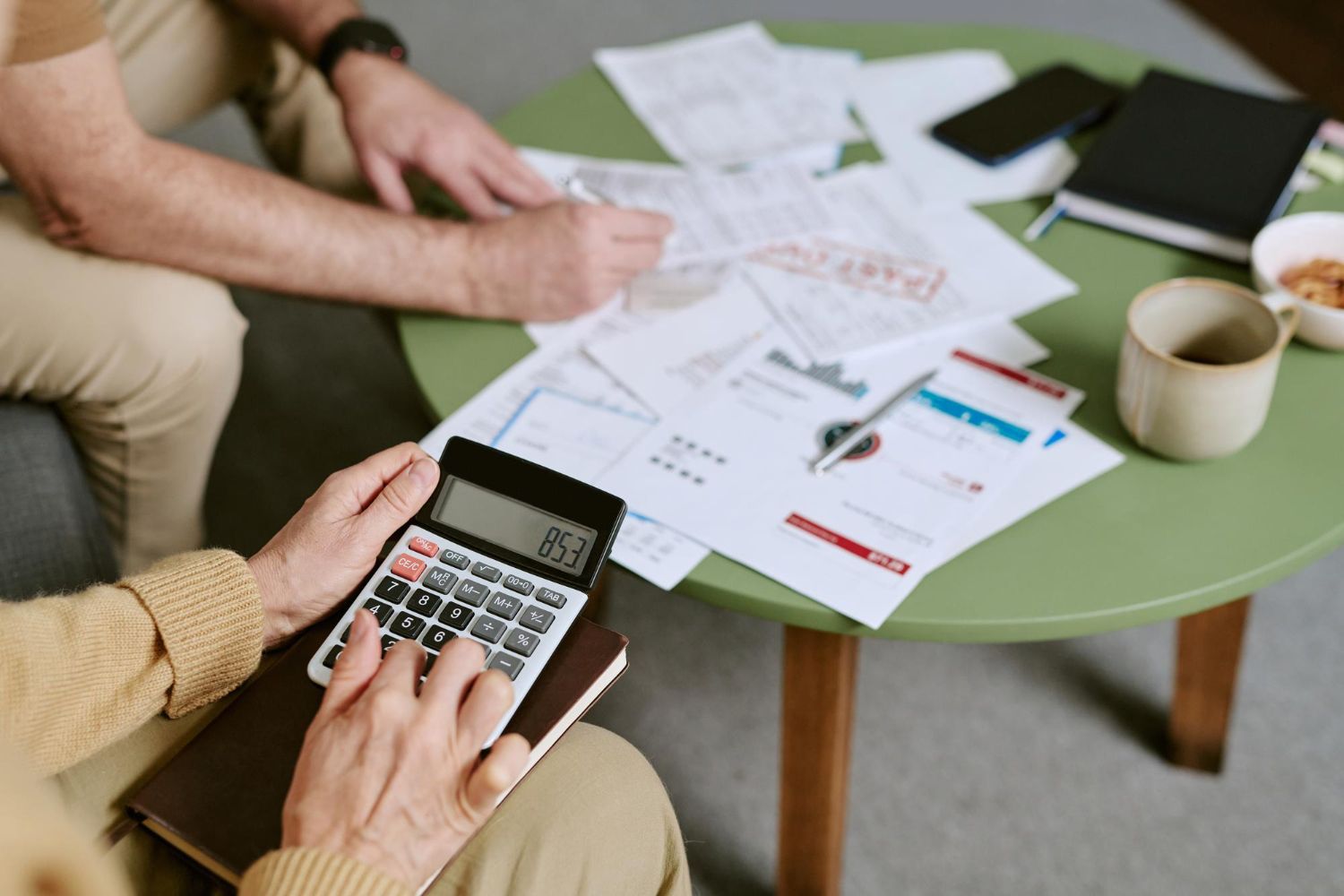 Two people reviewing printed charts and a calculator at a green table with coffee cup and notebook