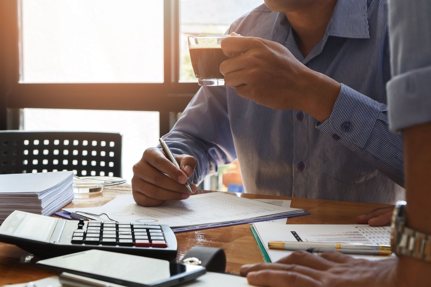 Colleagues reviewing documents and a calculator at a sunlit office desk, one holding a cup of tea