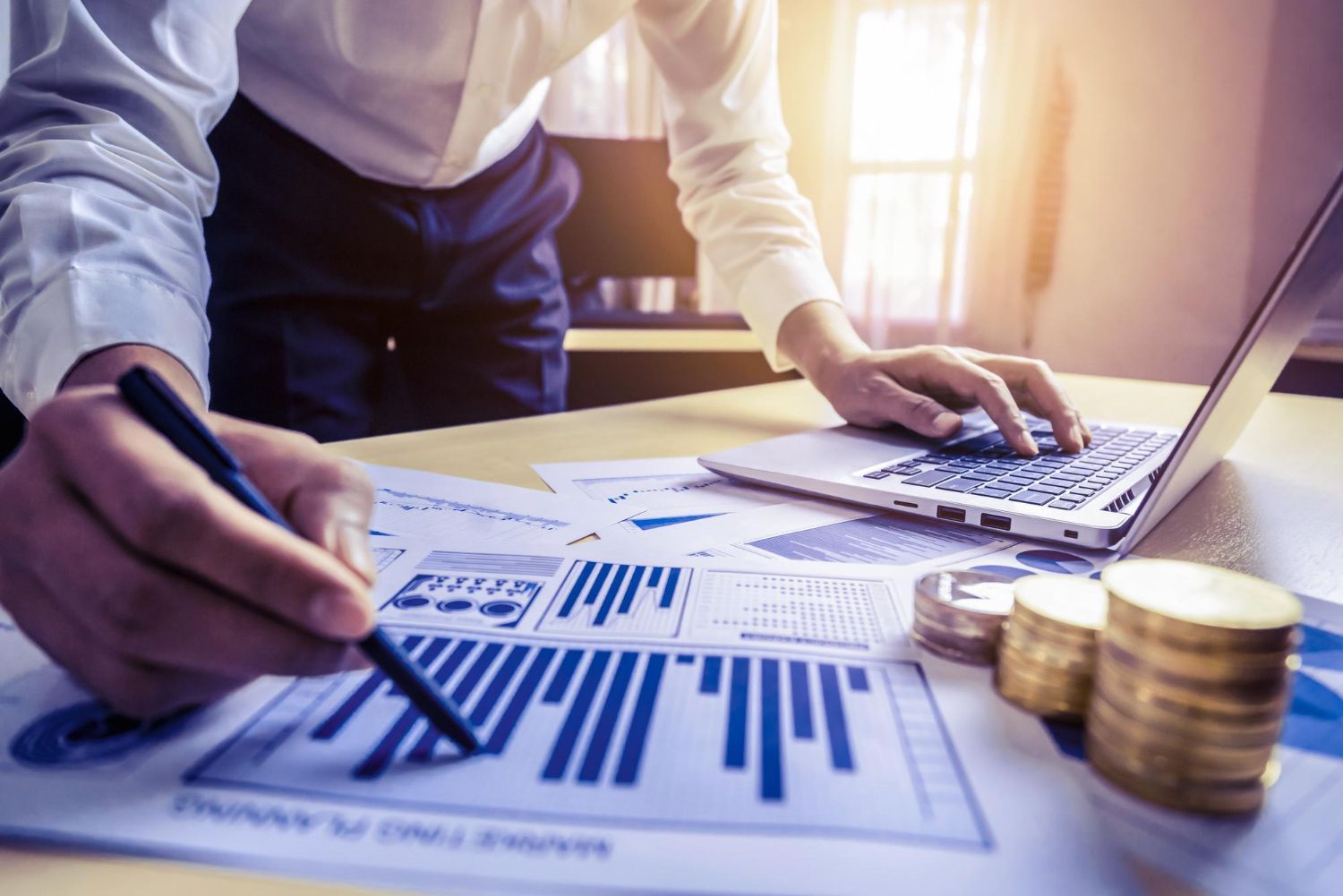 Hands marking financial charts beside a laptop and stacked coins on a desk, with warm sunlight.