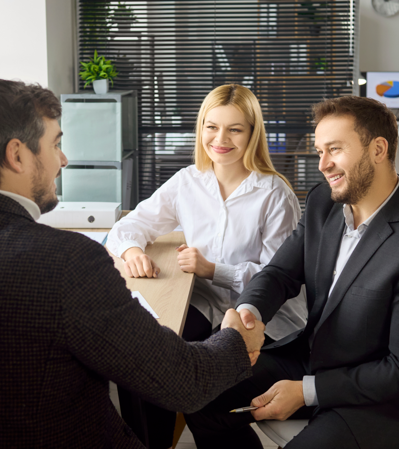 Business meeting with three colleagues shaking hands in an office, one smiling, others seated nearby.