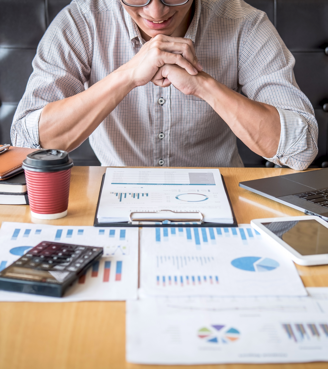 Person leaning over desk with charts, calculator, coffee cup, tablet, and laptop during financial planning.