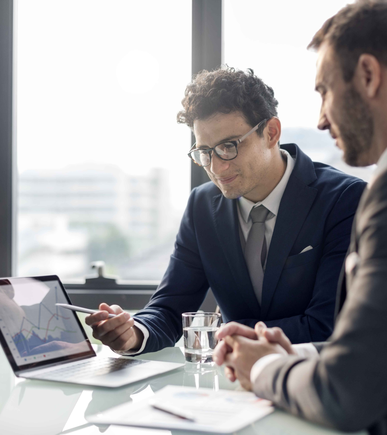 Two businesspeople in suits reviewing charts on a tablet during a meeting by a bright window