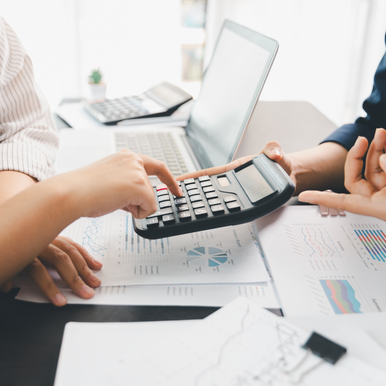 Hands using a calculator over financial charts at a desk with a laptop and papers