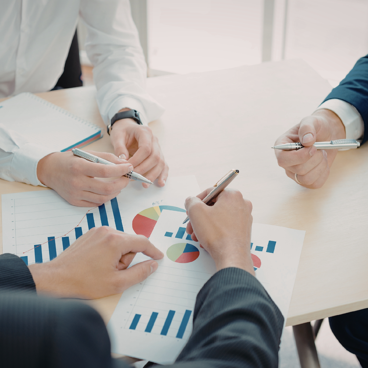 People in business attire reviewing charts and graphs around a table, pointing with pens during a meeting.