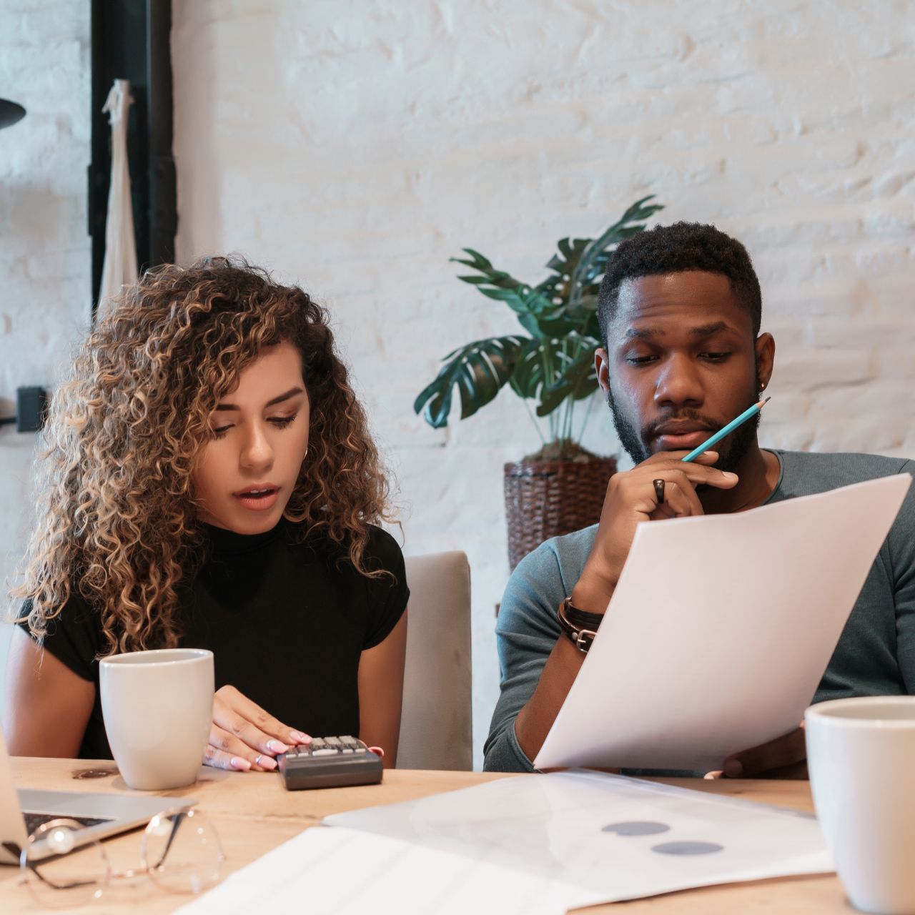 Two coworkers review documents at a table, one reading a laptop and the other taking notes.