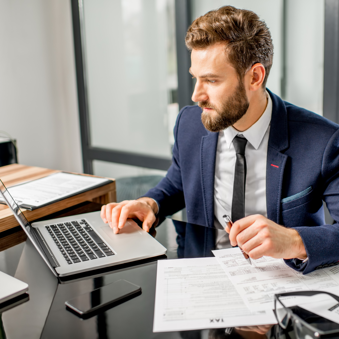 Man in a navy suit working at a desk with a laptop and papers in a bright office