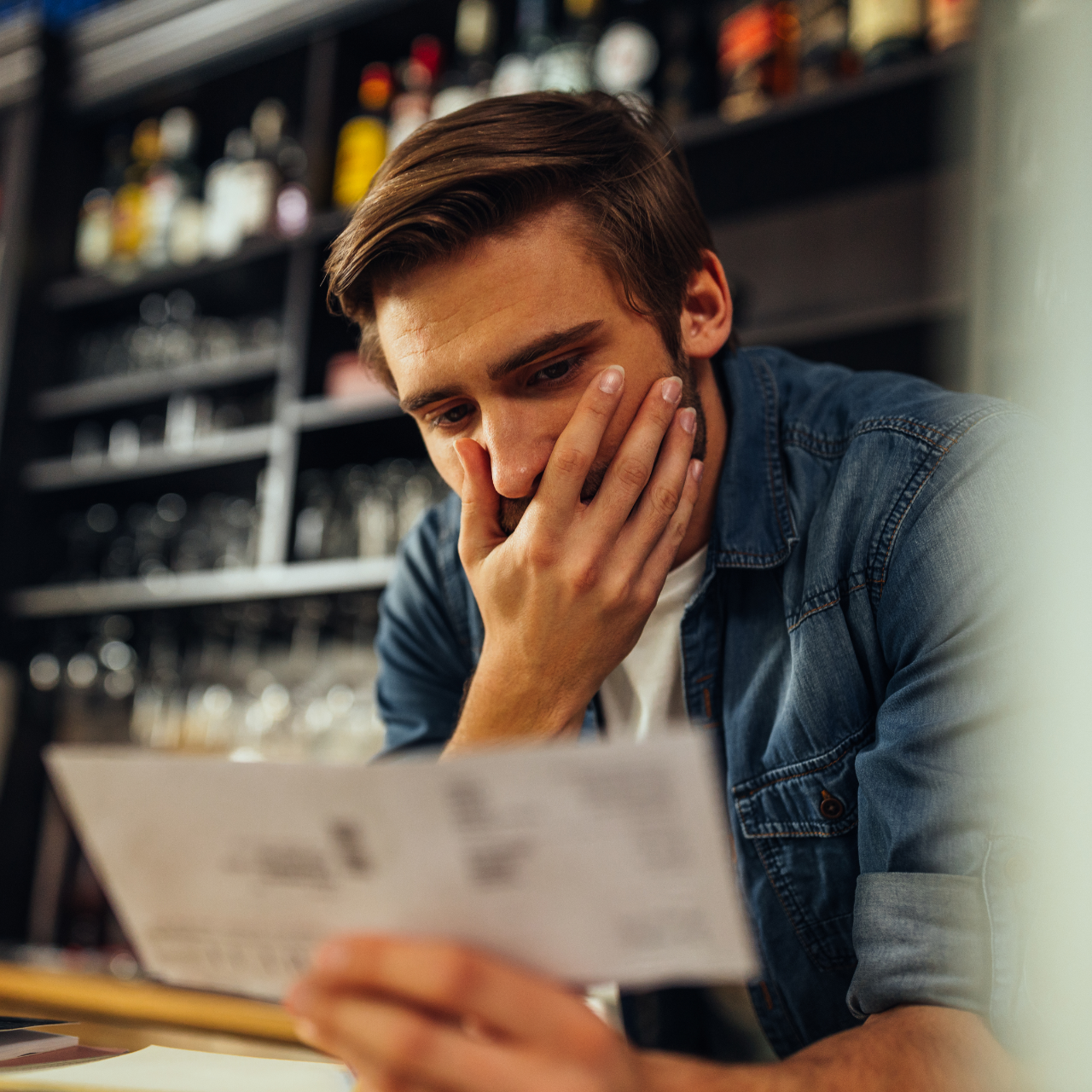 Person in a denim shirt reading a paper with a worried expression at a bar counter