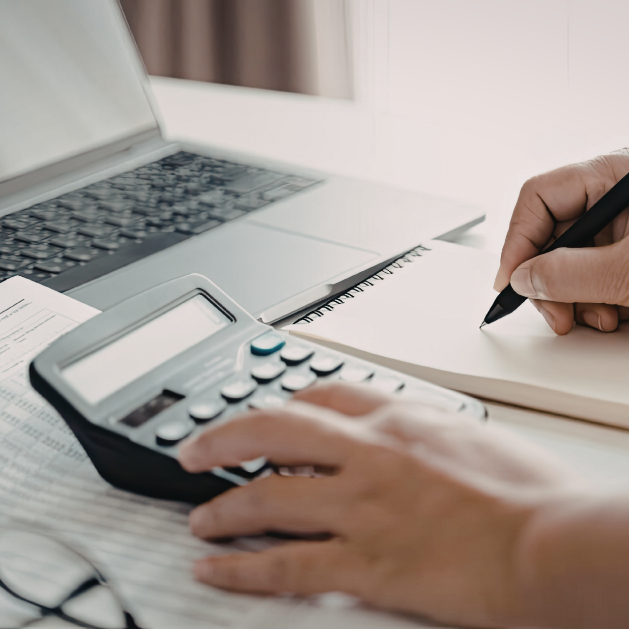 Hands calculating on a calculator beside a laptop while writing in a notebook
