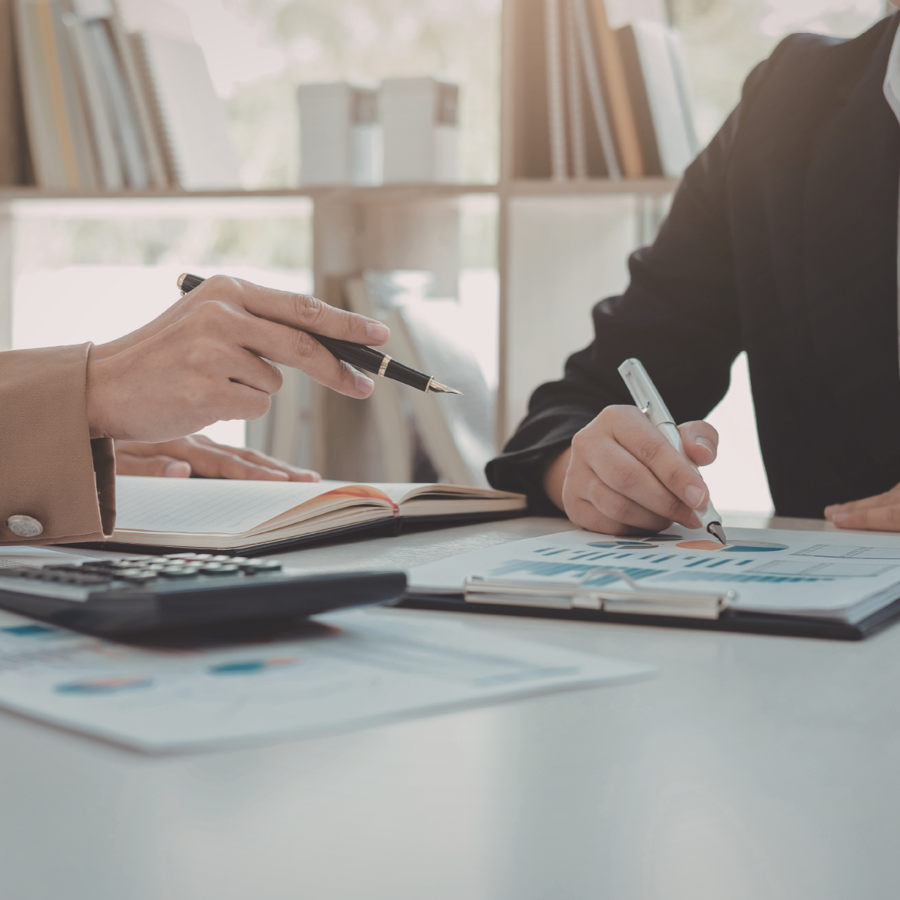 Two people reviewing documents and writing at a desk with charts, calculator, and pen in a bright office