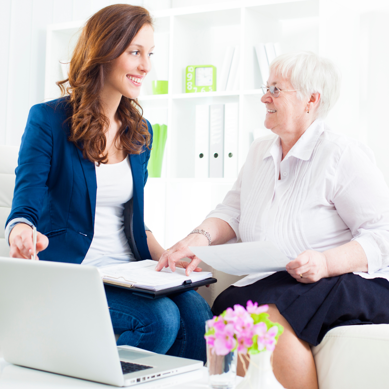 Two women talking in a bright office, one using a laptop while the other holds papers.