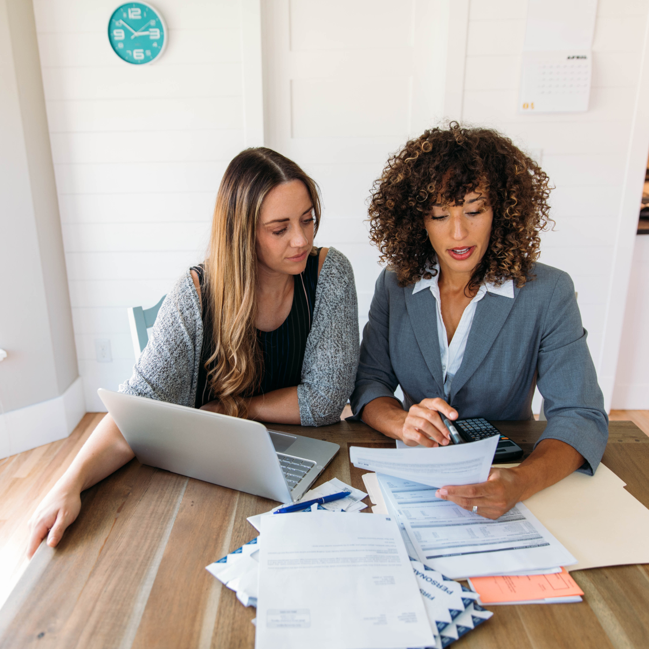 Two coworkers review paperwork at a laptop on a table in a bright office
