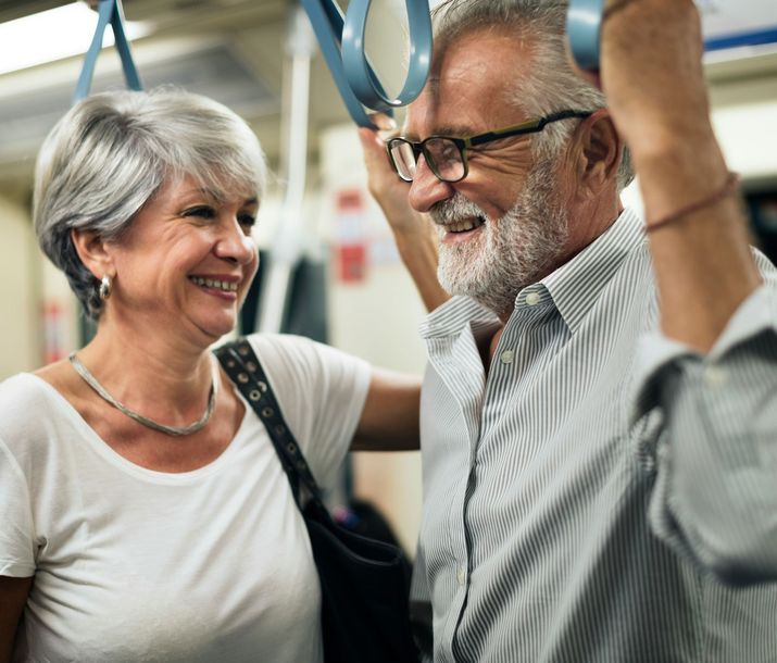 Two smiling people standing on a train, holding overhead straps, in a bright commuter car.