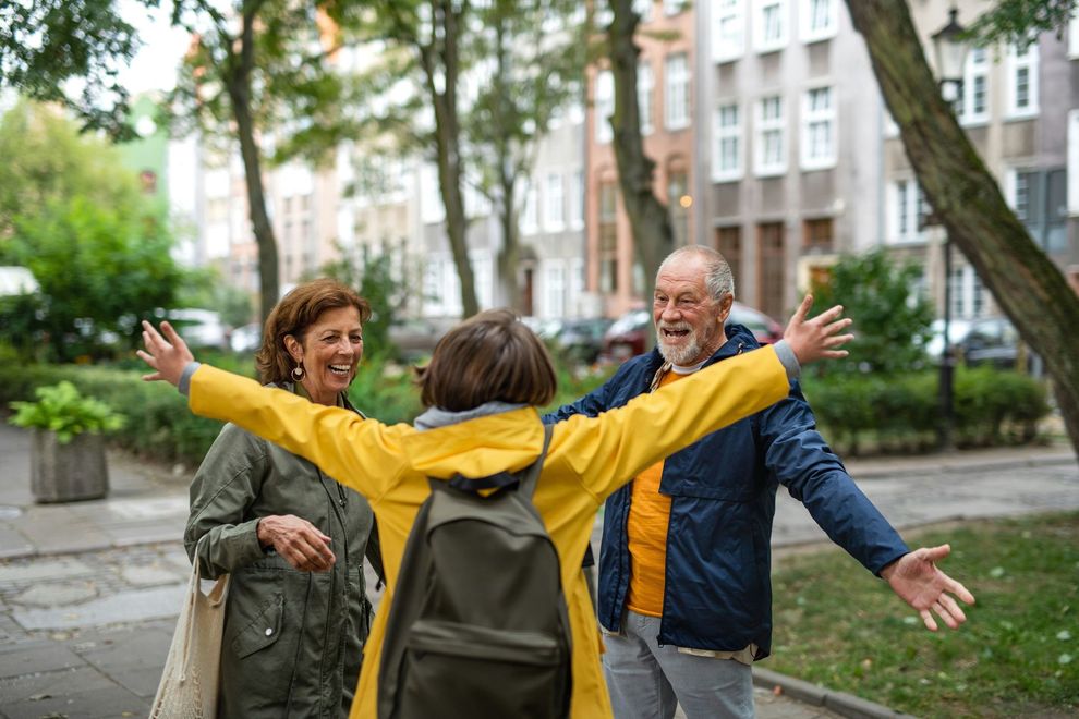 Three people smiling with arms open on a city sidewalk lined with trees and apartment buildings