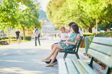 Woman and child sitting on a park bench in a sunlit park, with people walking in the background.