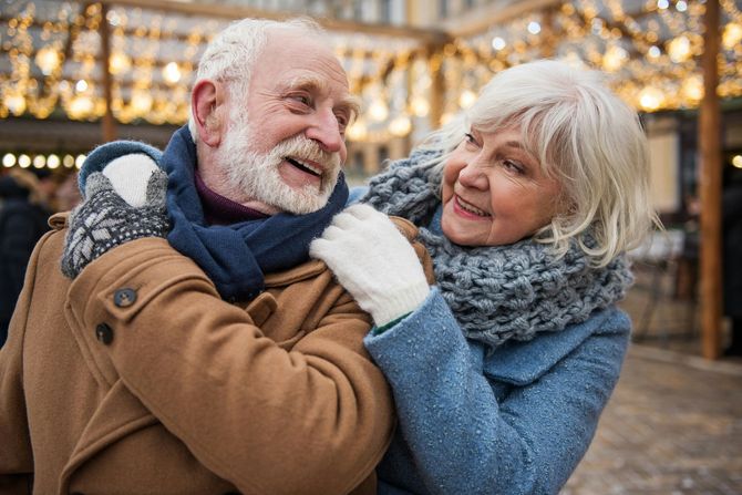Two smiling people in winter coats embrace outdoors under warm lights