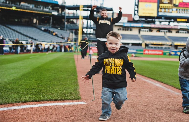 Child running on a baseball field near home plate, wearing a black shirt and jeans, with a stadium in the background