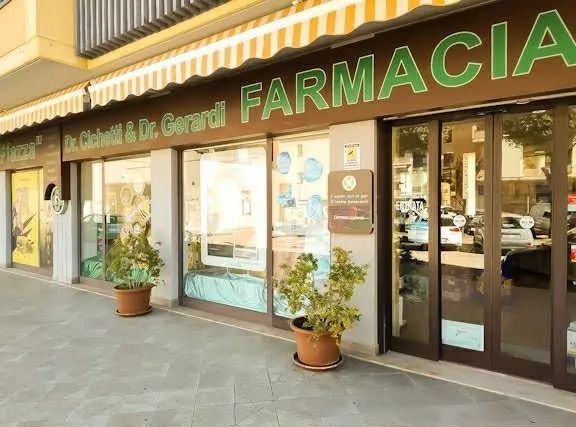 Negozio di farmacia a livello strada con insegna verde, vetrine e piante in vaso all'esterno.
