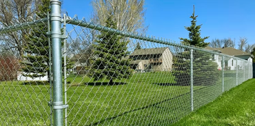 Chain-link fence along a grassy yard beside houses under a clear blue sky.