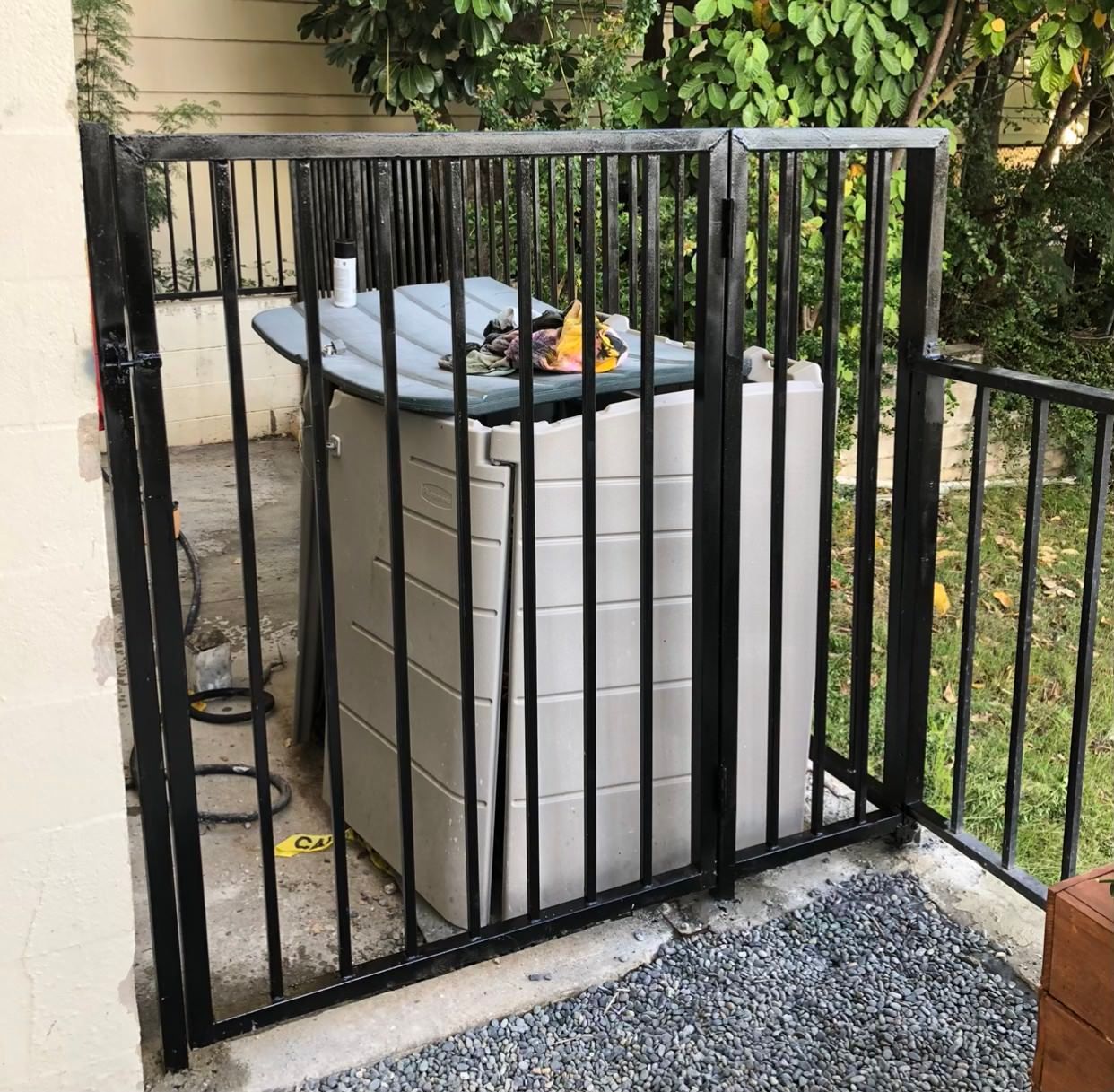 Black metal fenced enclosure around a large, tan plastic container; yard setting with gravel.