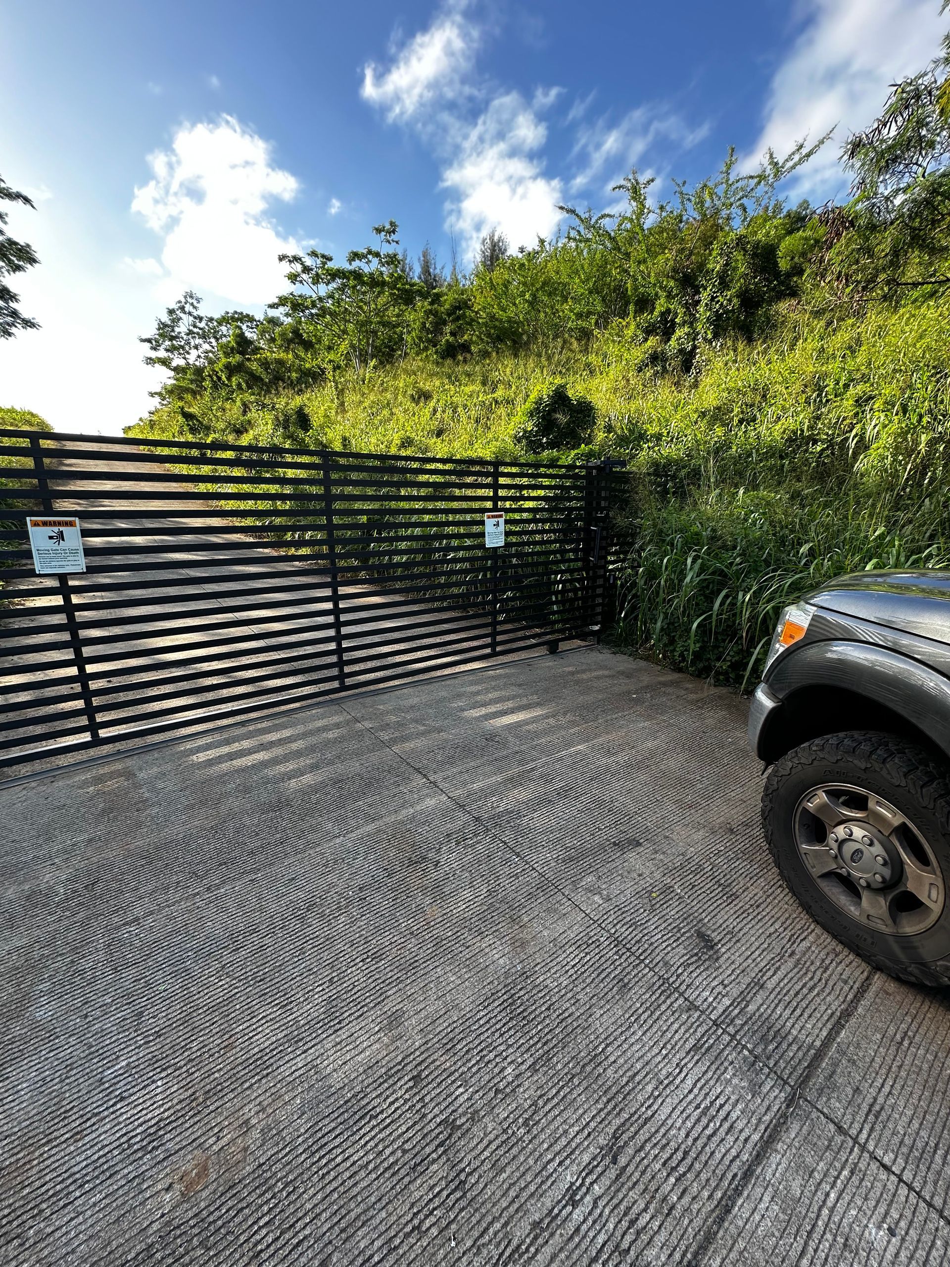 A dark gray vehicle parked on a patterned concrete driveway in front of a metal gate surrounded by green foliage.