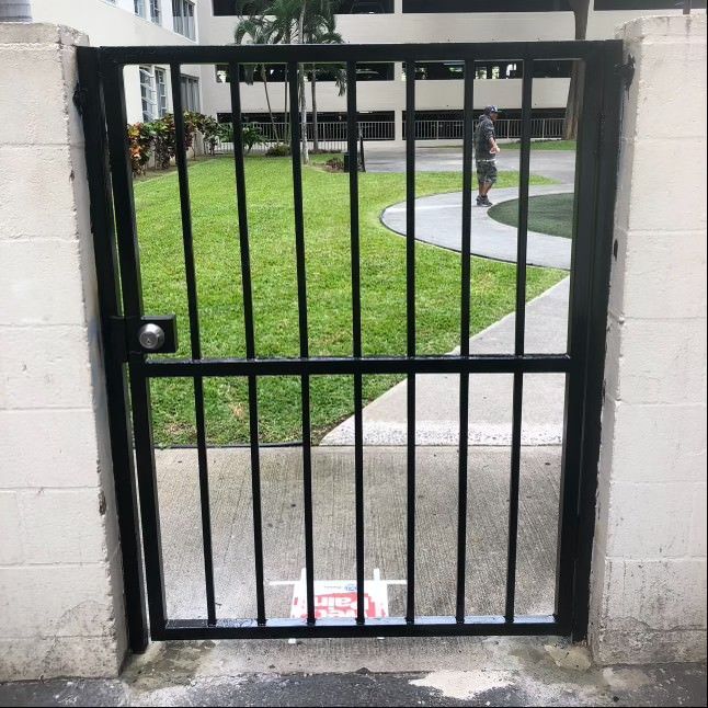 A black metal gate set in a cinder block wall, showing a sidewalk, green lawn, and person walking in the background.