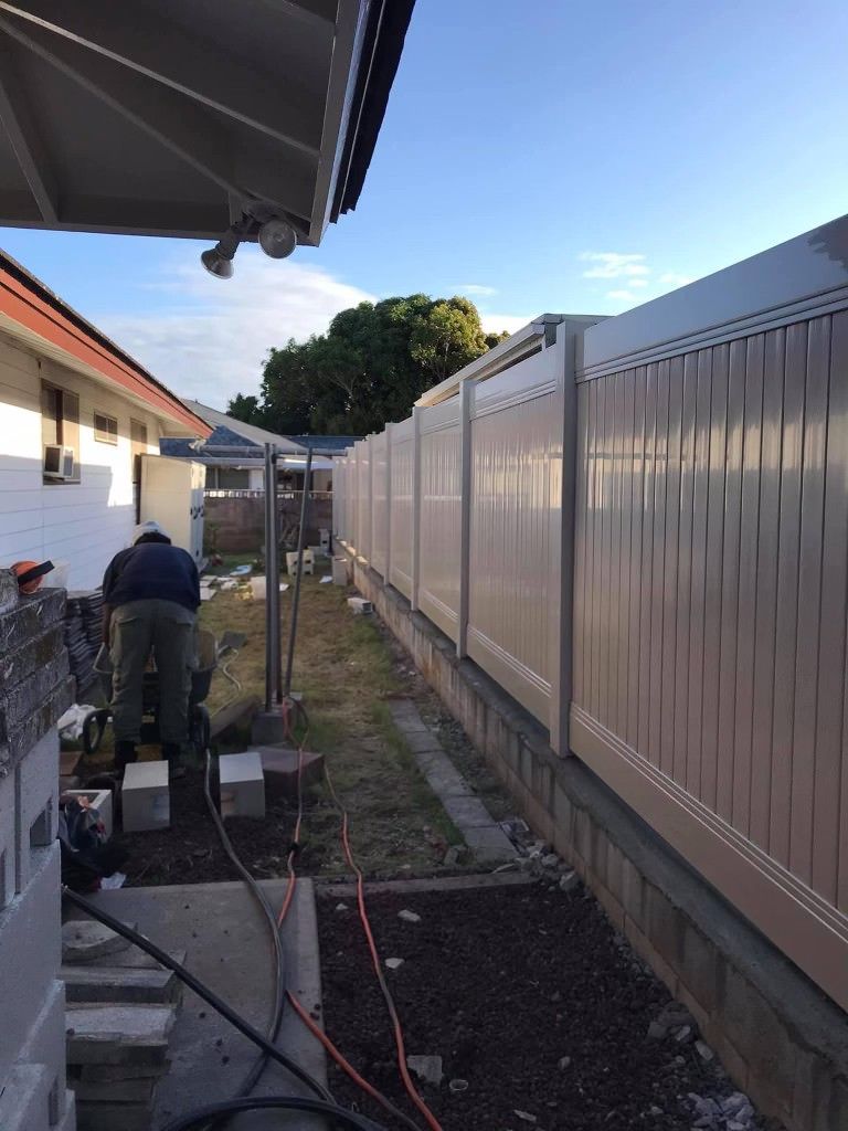 A person works in a side yard next to a newly installed tan vinyl fence mounted on a concrete base.