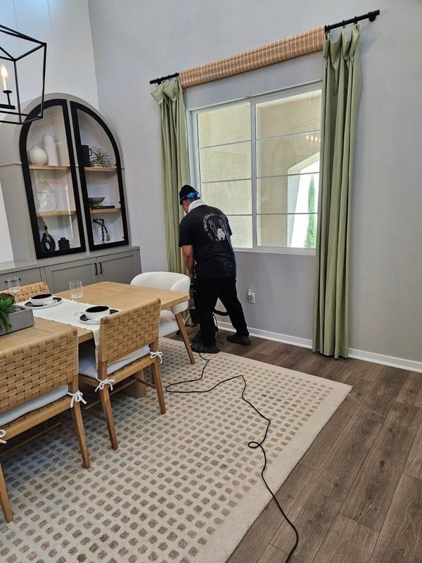 Person vacuuming a dining room with wicker chairs, a rug, and tall green curtains by a bright window