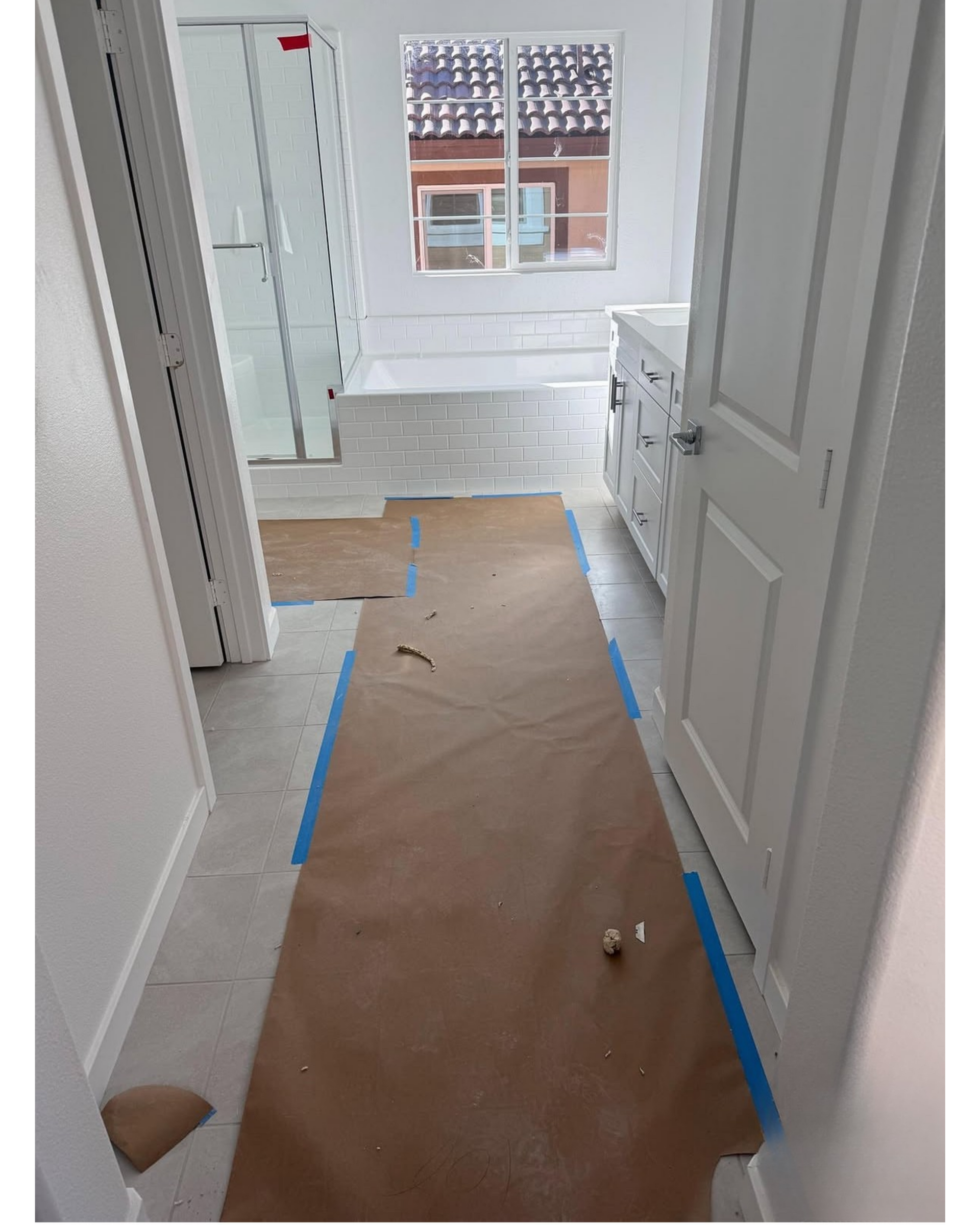 Hallway with brown floor covering taped down in blue, leading to a bright room with white doors and window.