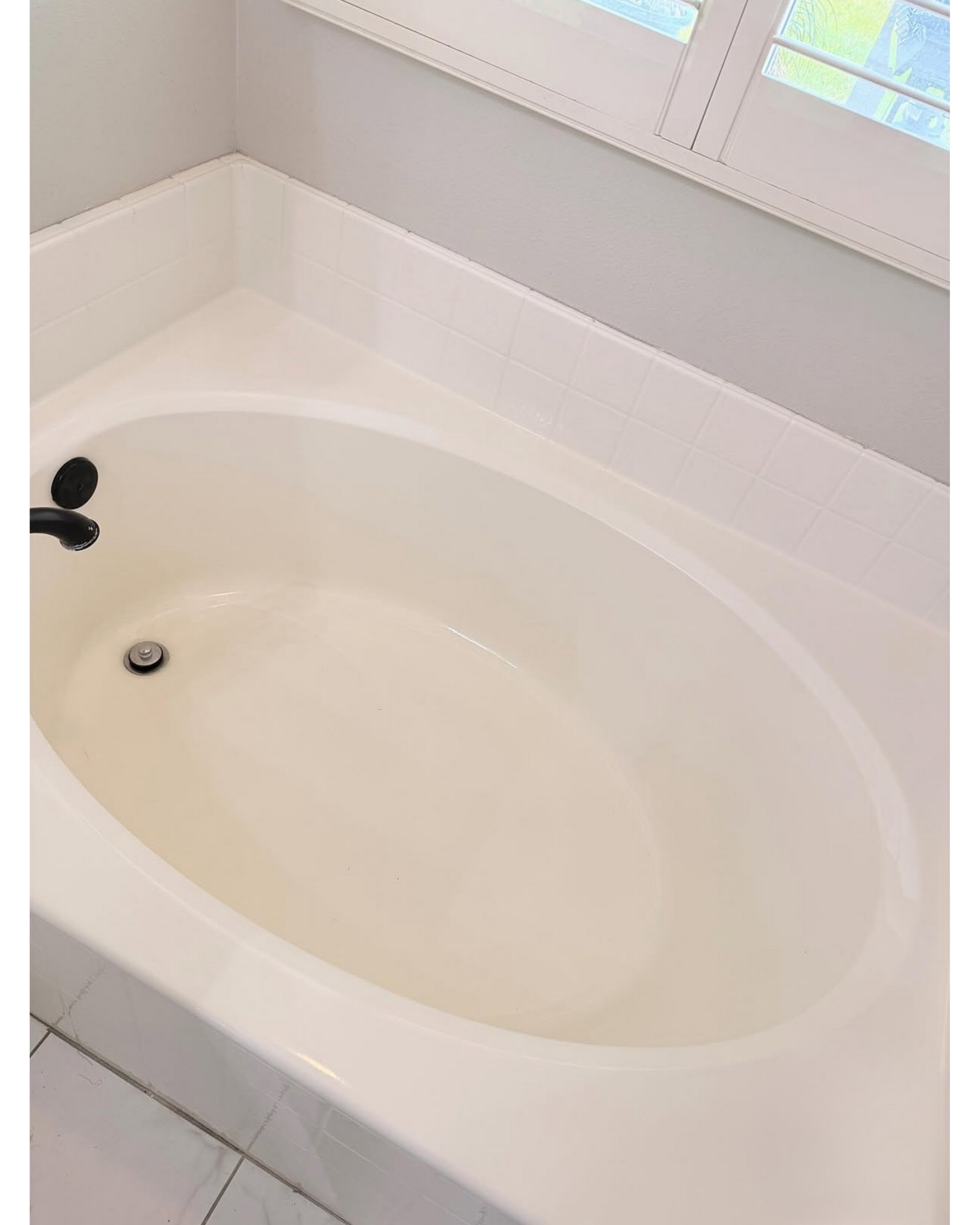 White oval bathtub with black faucet beside a window in a tiled bathroom