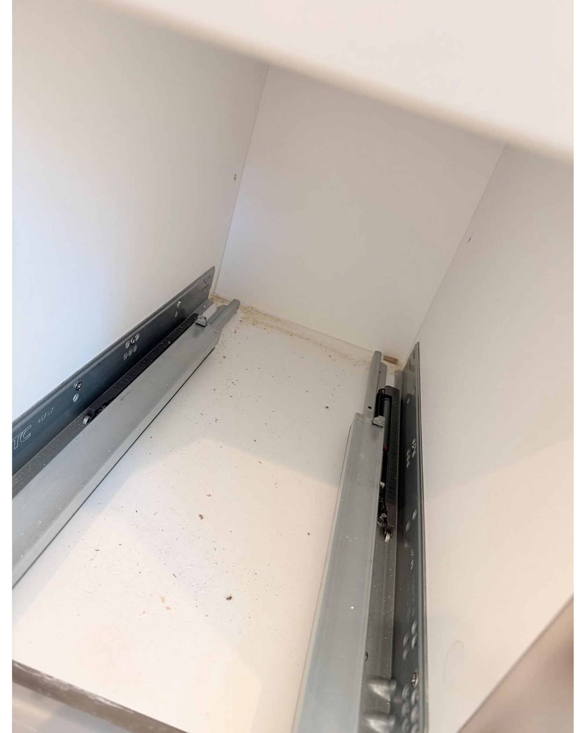 Empty white cabinet interior with metal drawer slides on both sides and small debris on the bottom shelf