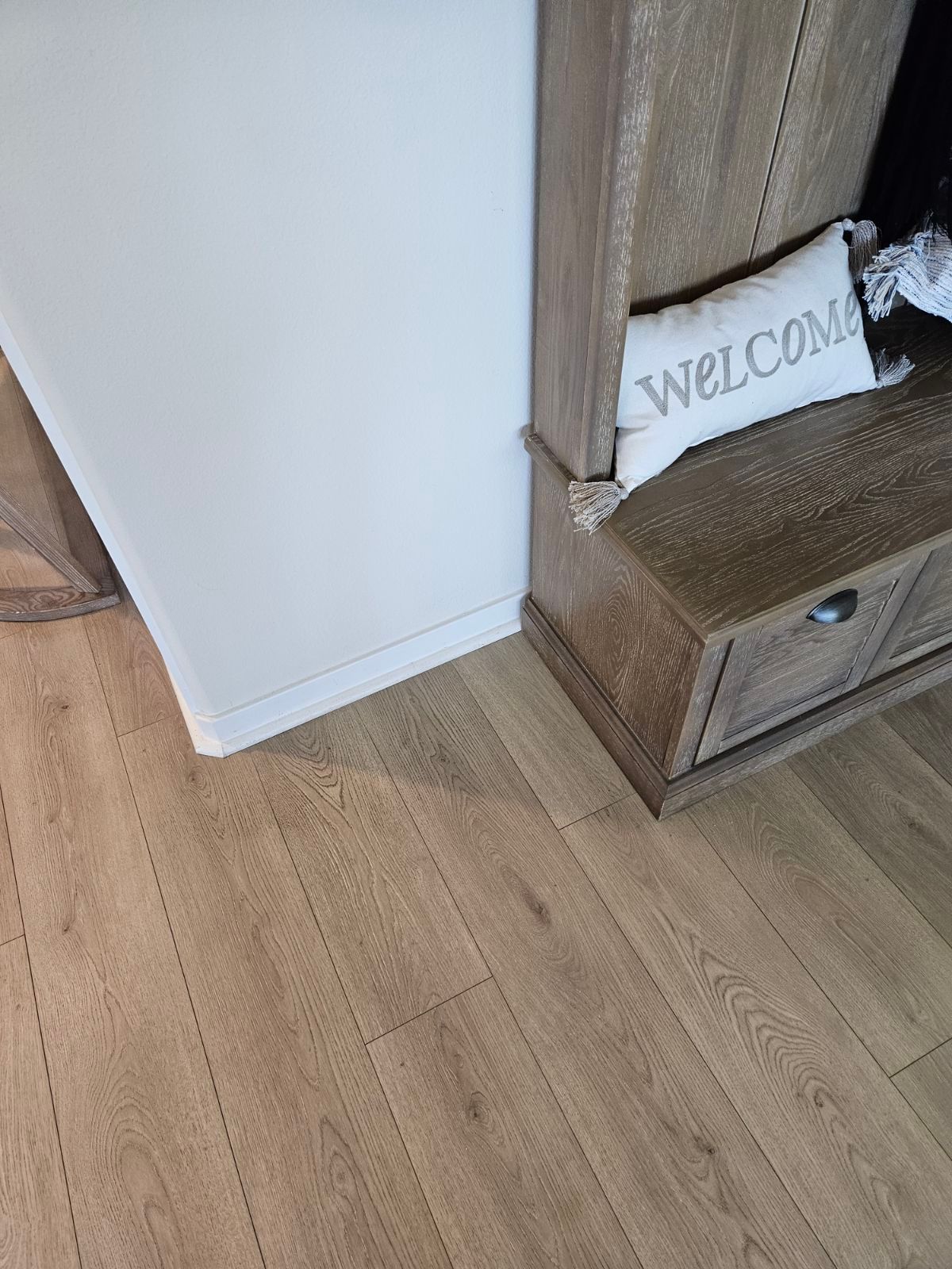 Corner of a room with light wood floor, white wall, and a wooden cabinet holding a “WELCOME” sign.