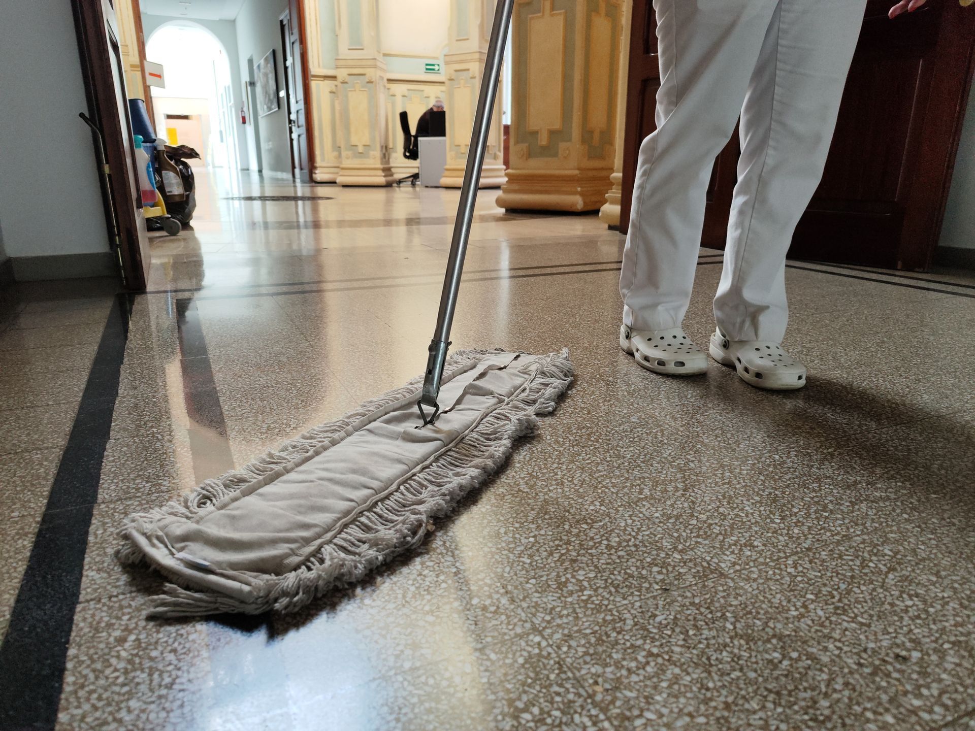 Person mopping a hallway floor with a gray mop in a bright indoor corridor.