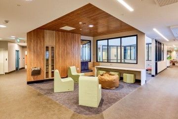 Modern lounge with wood-paneled seating nook, green chairs, table, and large windows in a bright office lobby