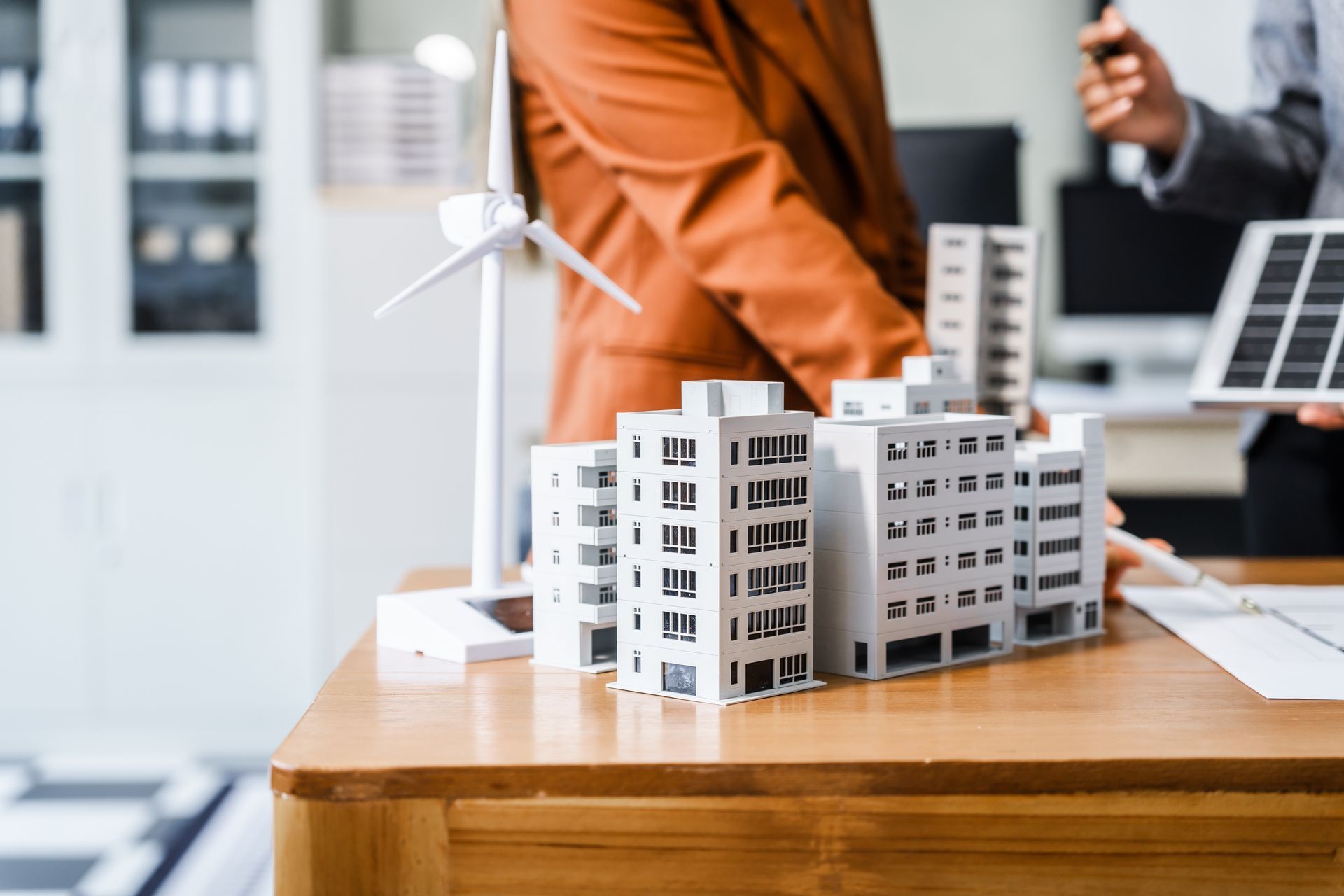 Miniature apartment buildings on a desk in a meeting room, with people and a laptop in the background Miniature apartment buildings on a desk in a meeting room, with people and a laptop in the background