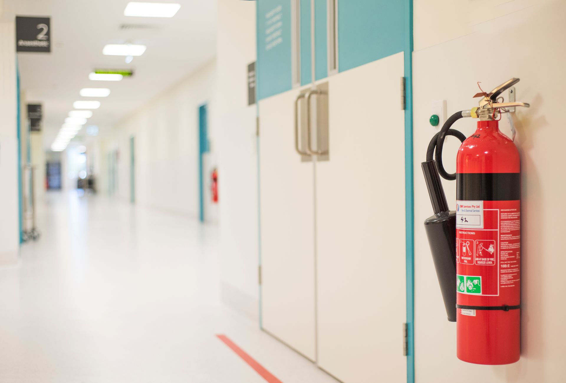 Hospital hallway with a red fire extinguisher mounted beside white cabinets and blue wall panels