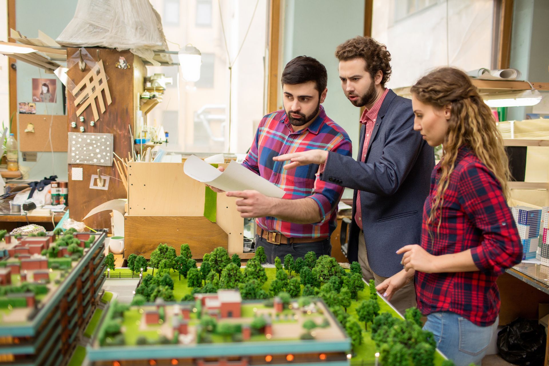 Three people reviewing a model landscape in a workshop, with miniature buildings and greenery on the table. Three people reviewing a model landscape in a workshop, with miniature buildings and greenery on the table.