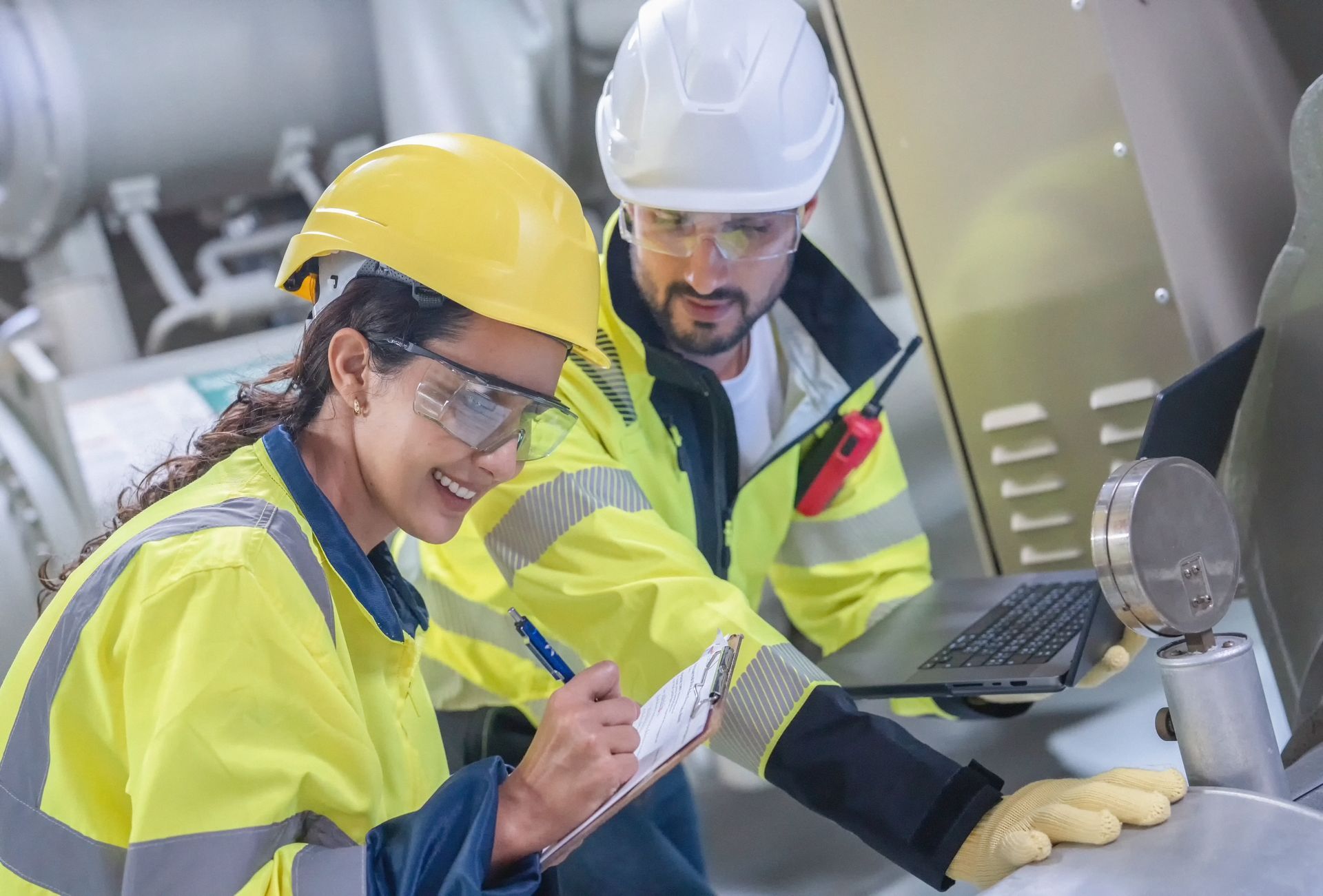 Two engineers in yellow safety gear inspect machinery in an industrial facility, one taking notes.