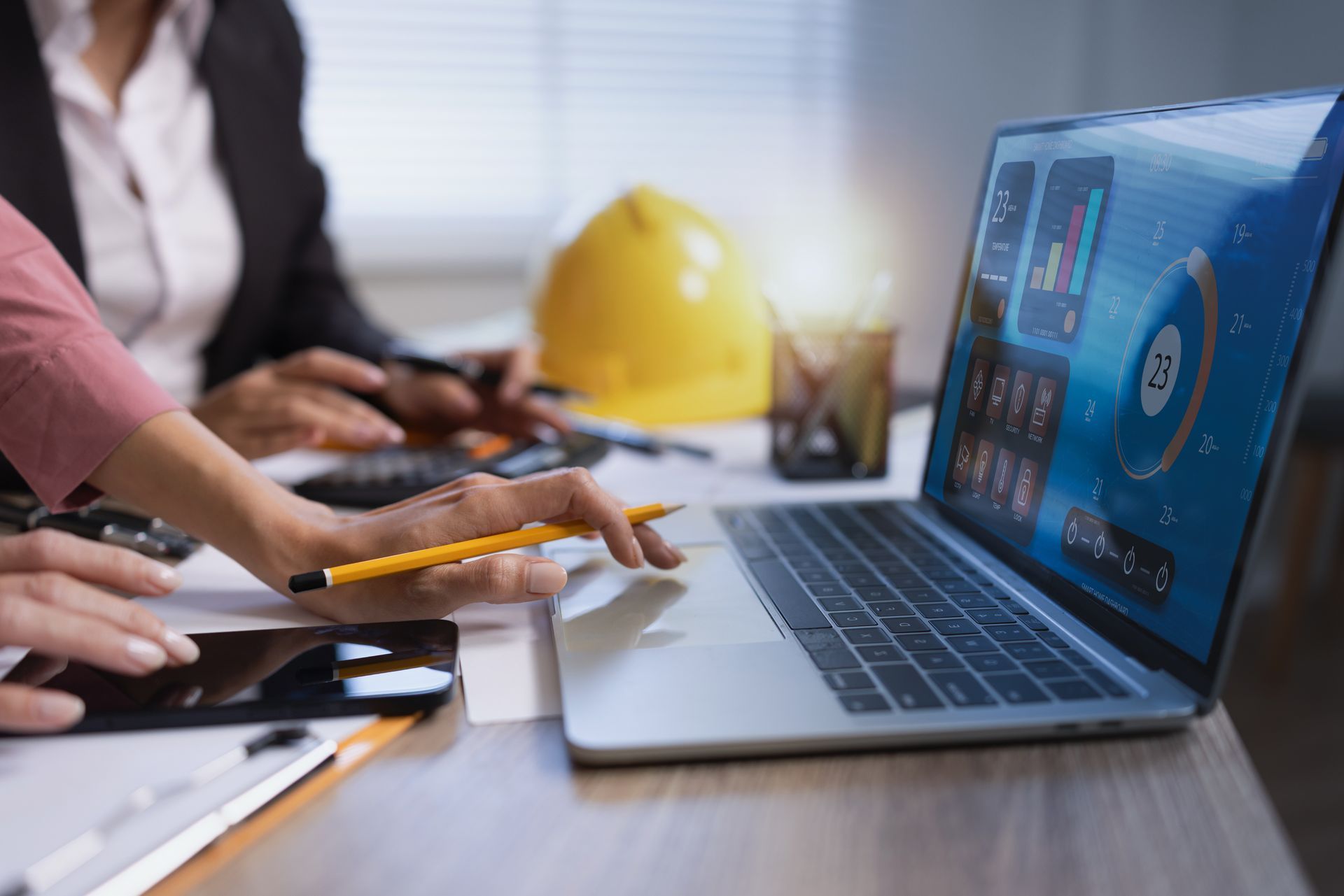 Team collaborating at a desk with a laptop, smartphone, and yellow hard hat in a meeting room Team collaborating at a desk with a laptop, smartphone, and yellow hard hat in a meeting room
