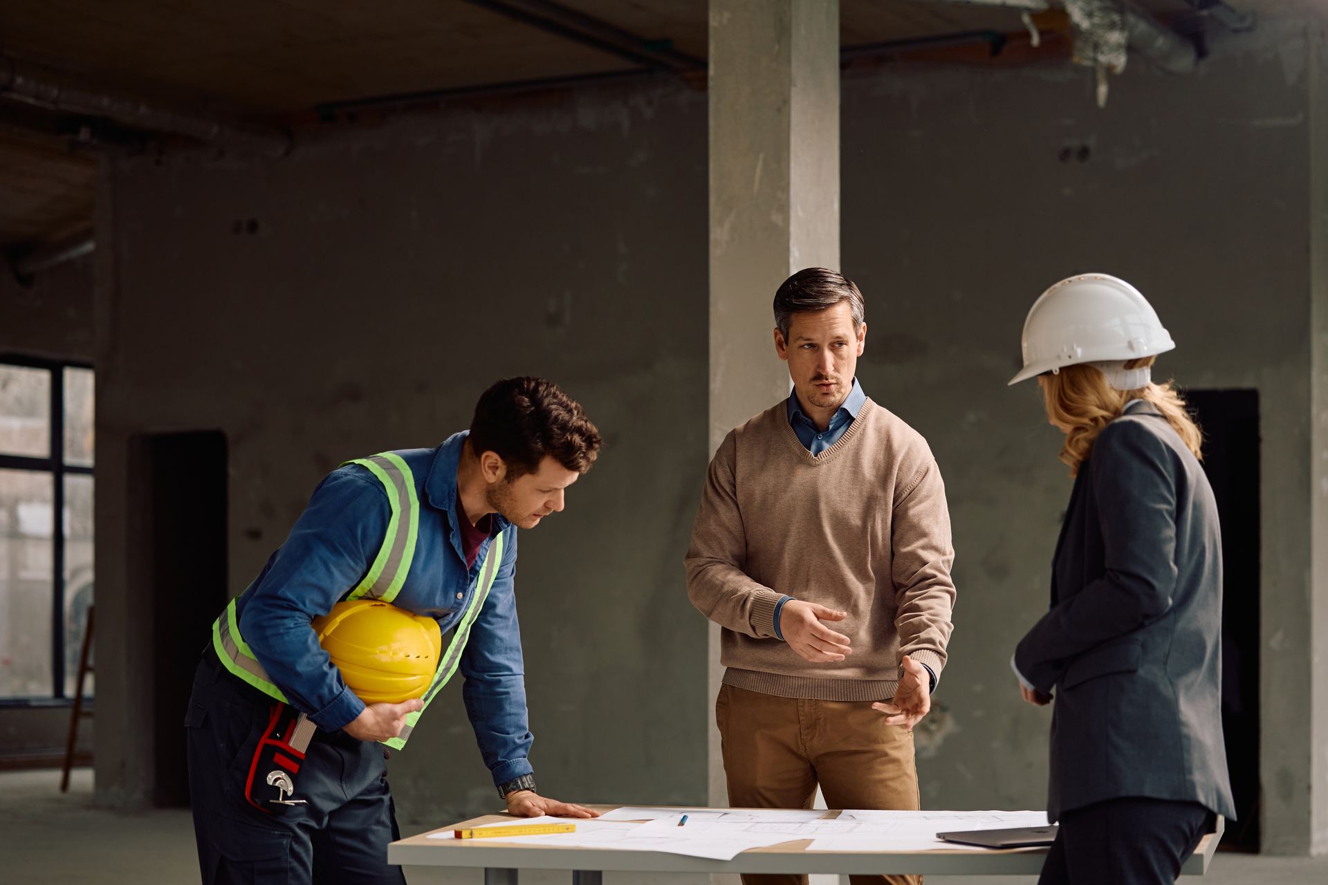 Three construction workers review blueprints at a worksite, one holding a yellow hard hat.