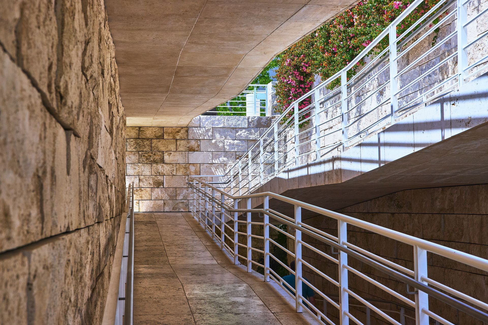 Narrow concrete corridor with metal railings and stone walls, leading into a bright outdoor walkway.