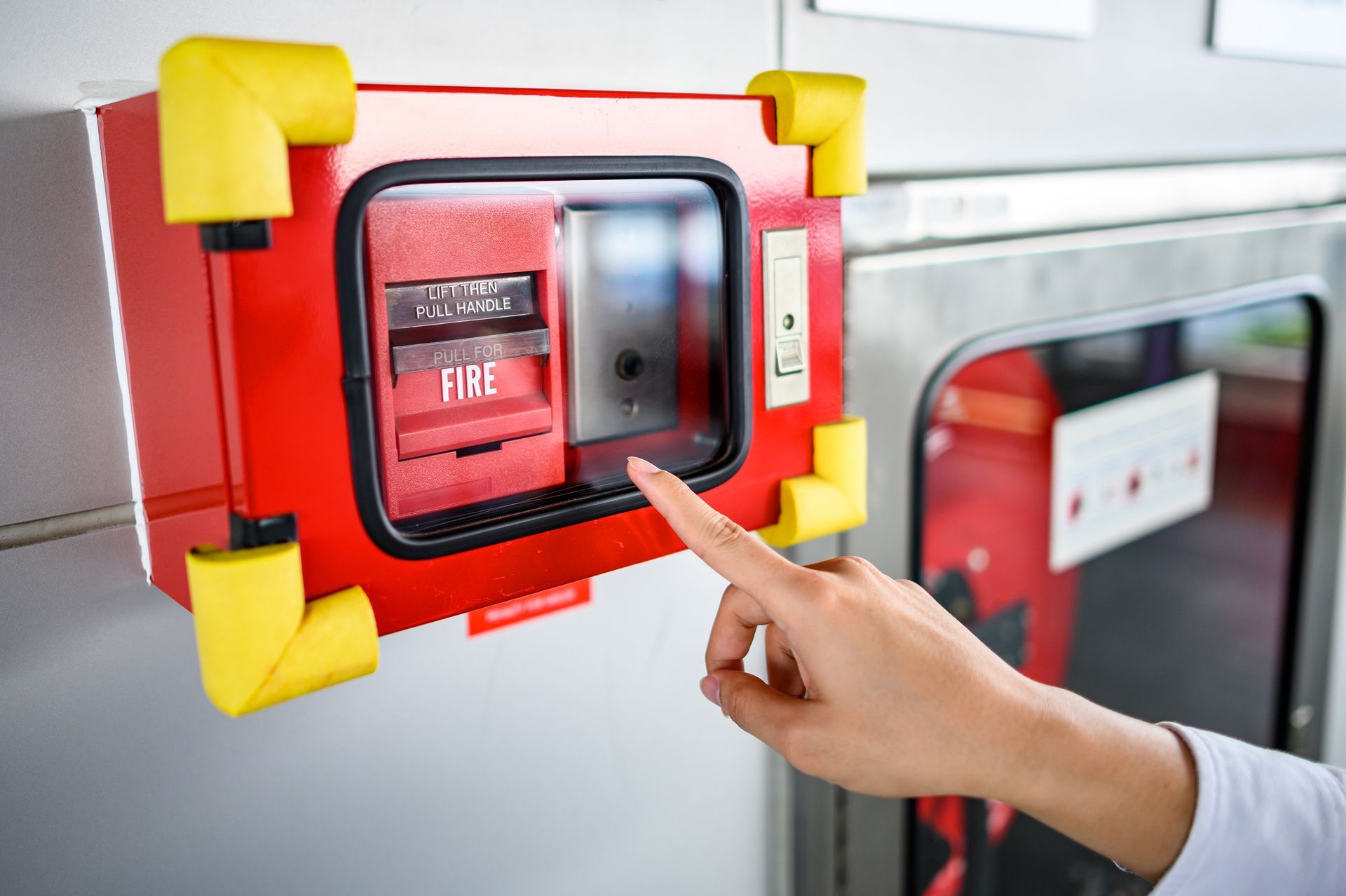 Hand pointing at a red fire alarm pull station with yellow corner guards on a wall in a transit area