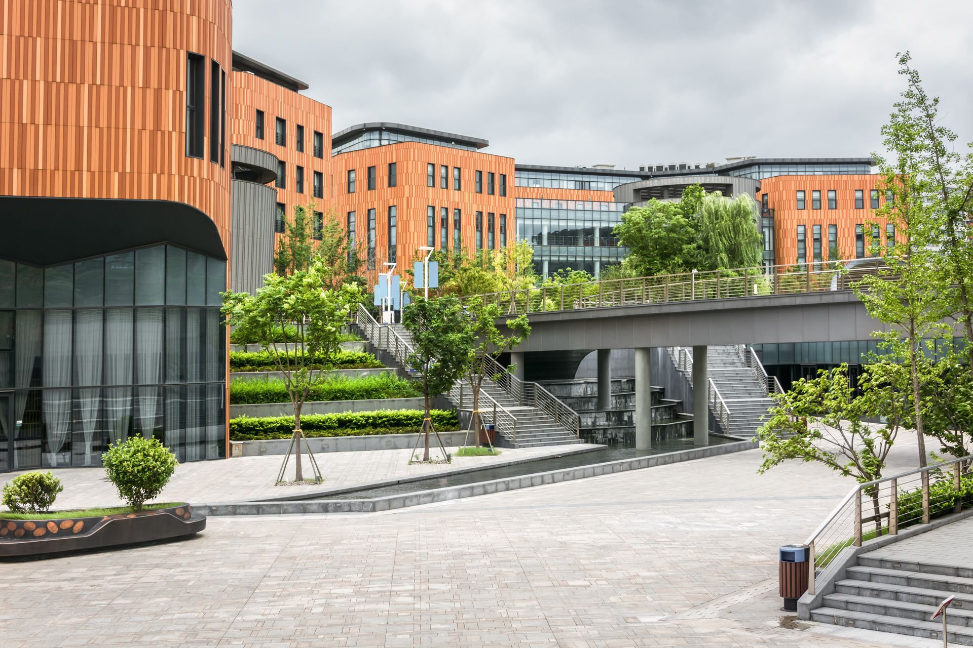 Modern campus courtyard with orange buildings, trees, and a pedestrian bridge over a paved plaza