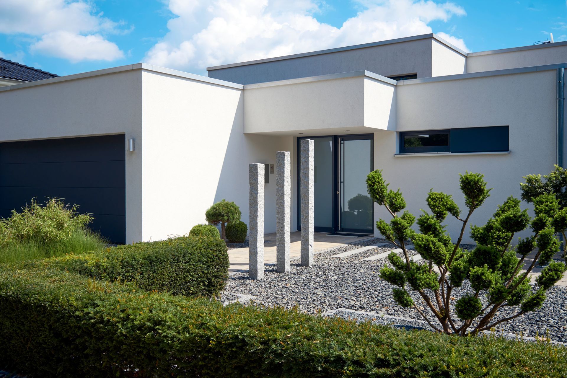 Modern white house with a dark garage door, front garden, and gravel path under a blue sky