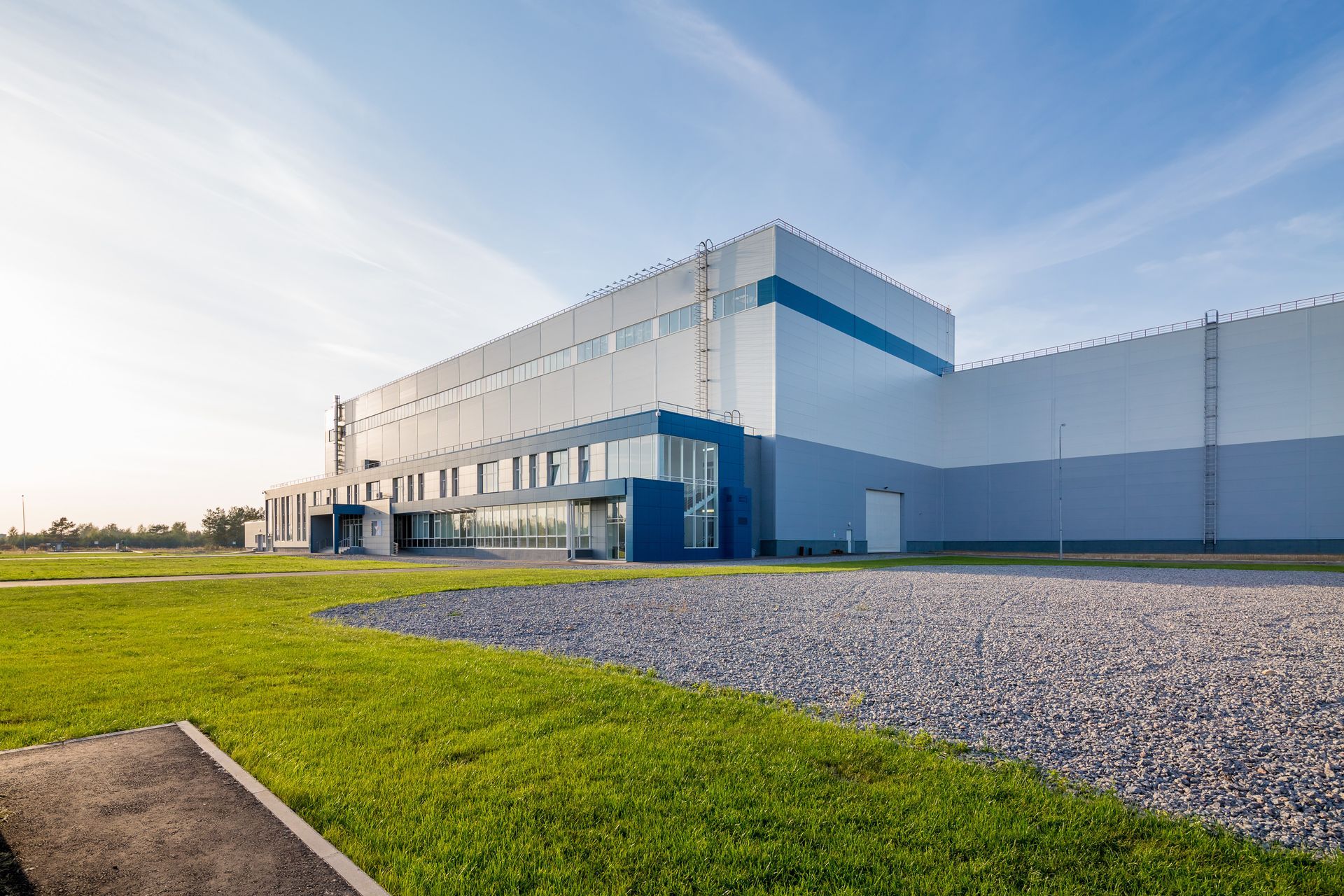 Modern white industrial building with blue accents beside a grassy field and gravel lot under a clear sky Modern white industrial building with blue accents beside a grassy field and gravel lot under a clear sky