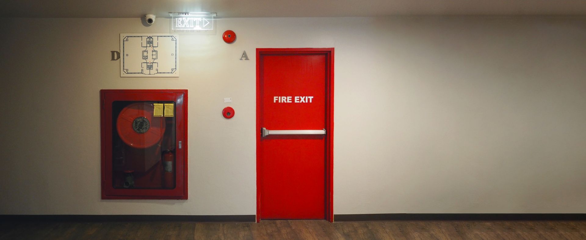 Red fire door and wall-mounted fire hose cabinet in a hallway with beige walls and wood floor