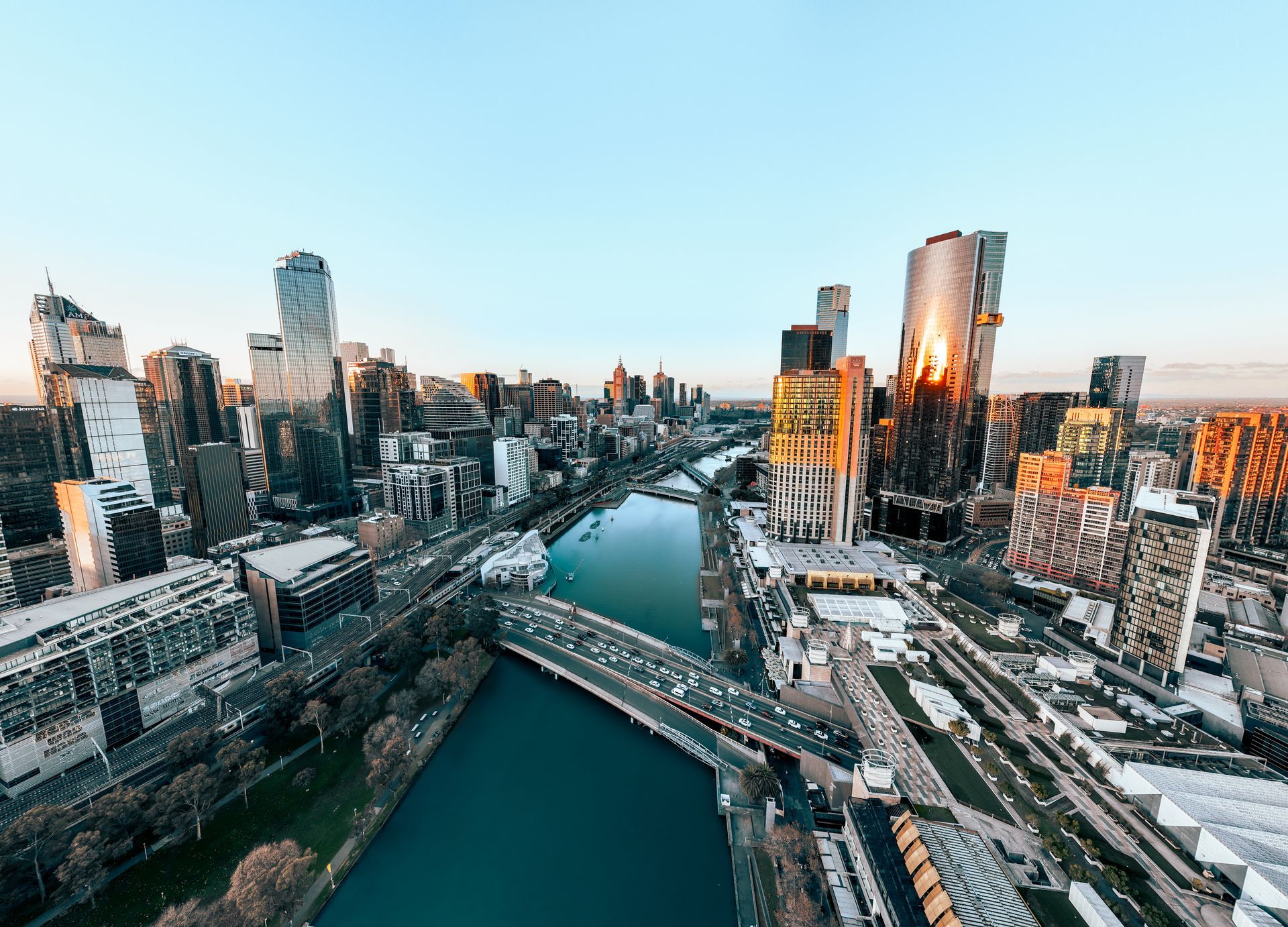 Aerial view of a city skyline with a river and bridge at sunset, buildings reflecting warm light.