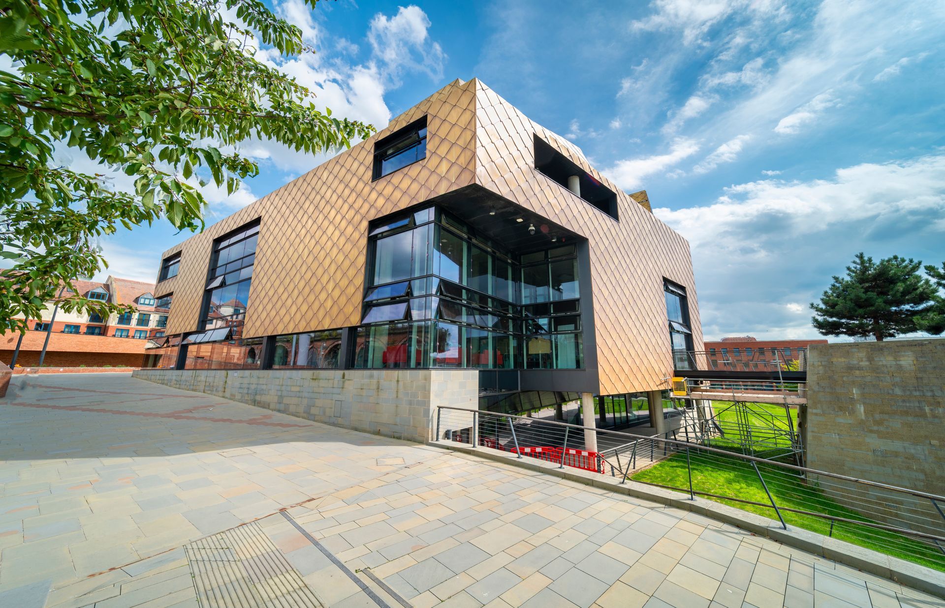 Modern brick-and-glass building with a paved courtyard and trees under a blue sky