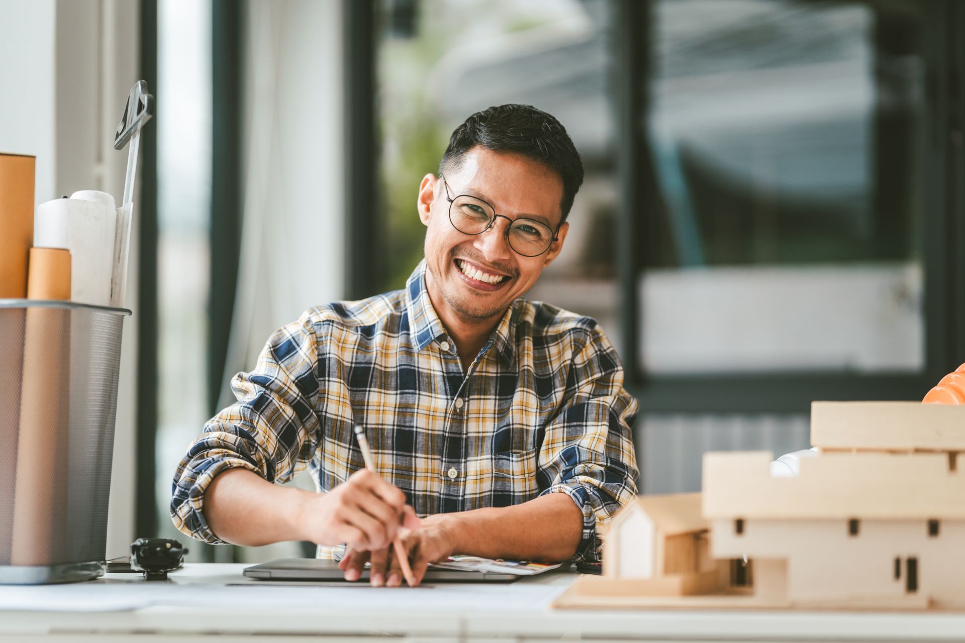 Smiling person sketching at a desk beside architectural model in a bright studio